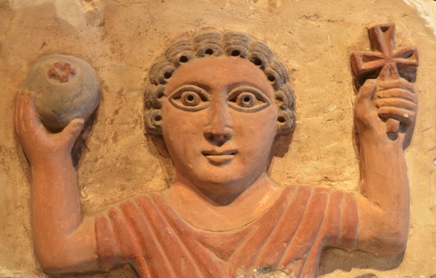 antique stone carving (relief) of a man with curly hair, both hands raised, one hand holding a spherical object and the other hand holding a cross