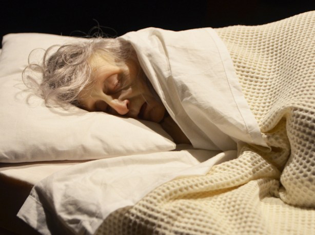very realistic and life like scupture of an old woman with grey asleep under a blanket with her head on a pillow