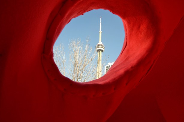 the CN tower with bright blue sky, as seen through the hole in a sculpture, the eye of a large red bear. 