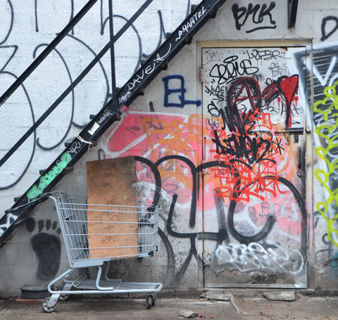 metal staircase, outside, running diagonally across the back of a building, a shopping cart under the stairs with a box in in, a door in the wall under the stairs, both door and wall are cover in graffiti including a large orange swath and a bright red heart 