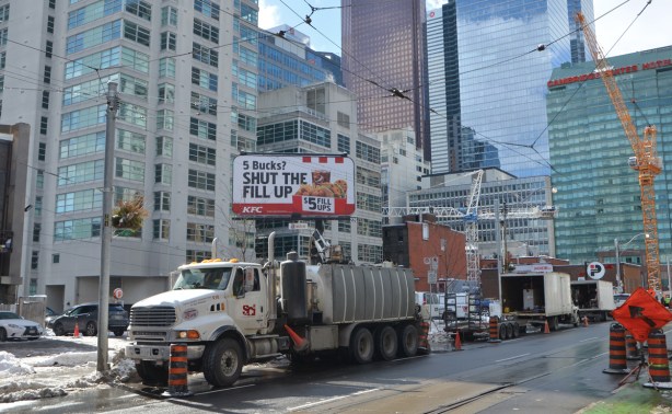 large truck parked on a street with tall buildings behind, and a large billboard with a KFC ad on it 
