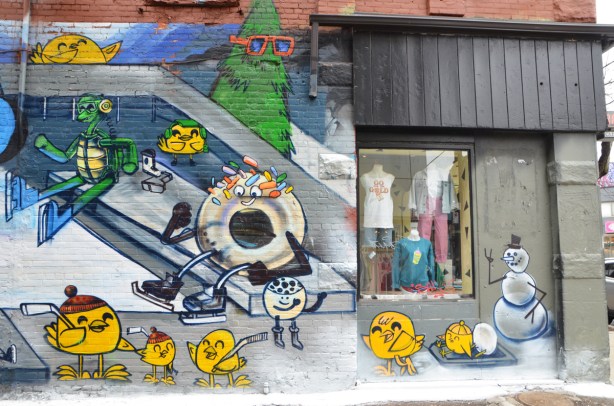 right hand side of a large mural by uber5000 in Graffiti alley, with shop window in the picture.  Theme of the mural is skating at Nathan Phillips square in toronto.  little yellow birdies, a green frog is puttin on skates as is a donut with white frosting and a few sprinkles