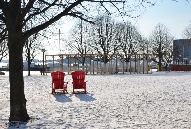 the back side of two red muskoka chairs in a snow covered park with a large art installation of wind chimes in the background 