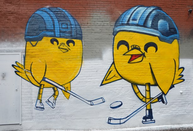 two large yellow uber5000 birdies with hockey sticks and old fashioned hockey helmets. One is wearing white skates and the other is wearing black skates. mural in graffiti alley 