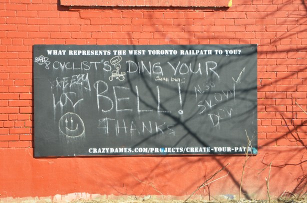 large chalkboard on an orange brick wall with notes to tell cyclists to slow down and ring their bells.