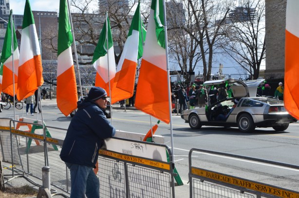 a man stands on a sidewalk, leaning on a barricade, lots of large Irish flags, a delorean car with its door open is across the street 