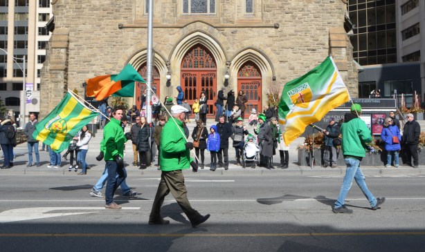 three men carrying flags in the St. Patricks day parade, walk past a church, many people on the sidewalk, pavement, watching the parade go by 