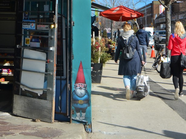 a small gnome painted on the wall beside a door to a convenience store. The door is open and people are walking past