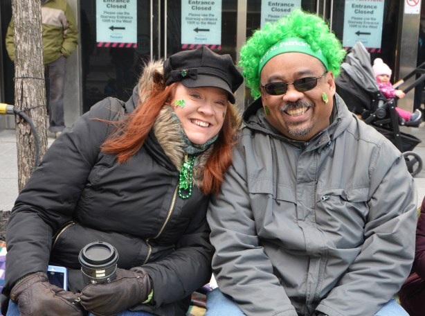 two people sitting on the sidewalk watching the St. Patricks Day parade, a red head woman with long hair and a man with a curly bright green wig, both are smiling, both dressed for cold weather, 