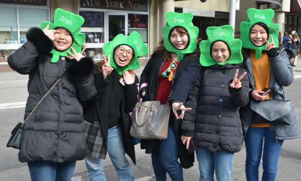 5 Asian women (Korean?) watching St. Patricks day parade, posing for the camera, all with shamrock green things aon their heads so only their faces stick out, the word Irish is written on each shamrock 