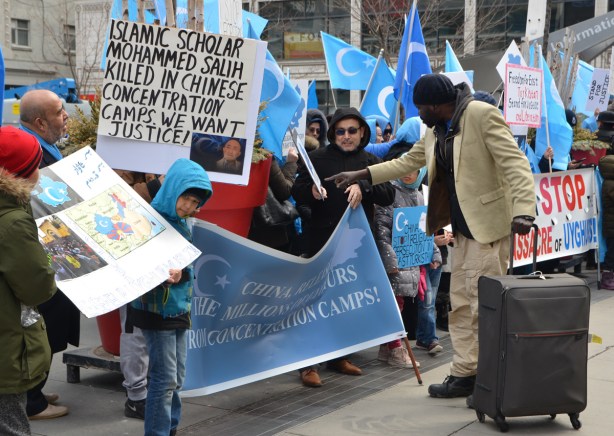 a black man with a suitcase has stopped to talk to people protesting for a free East Turkistan, he is pointing to one of the signs and a man is explaining something to him 