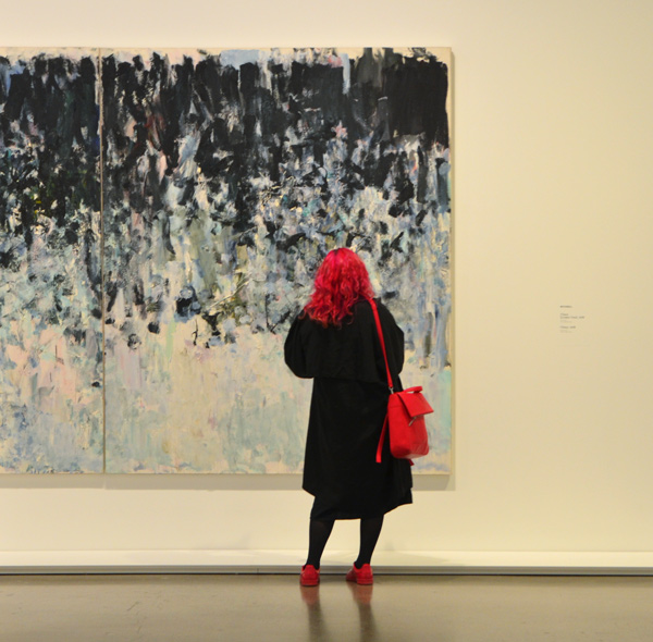A woman in dark bright pink hair, with matching purse and shoes stands in front of a large painting by Joan Mitchell in an art gallery 