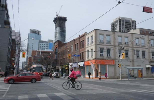 the intersection of Queen and Jarvis, looking northwest, with a foodora cyclist in pink in the intersection, traffic, street car, and in the distance, development on Yonge and westward, cranes, glass towers