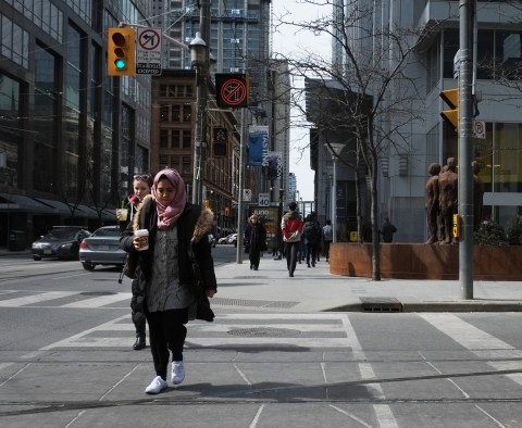 two women holding cups of coffee walk across Victoria St. on Queen Street East