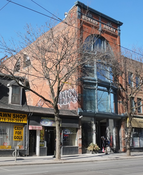 tall brick building, Richard Bigley 1876 written in white at the top (4 storeys), large glass windows in front, once an old store and warehouse, now a 3 loft apartments. 98 Queen Street East