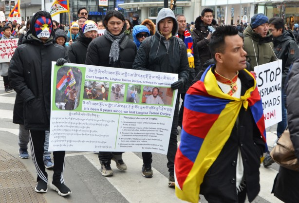a group of young men marching in a protest, Free tibet. carrying a banner with a lot of words in both Tibetan and English, one is wrapped in a Tibetan flag, some are wearing free tibet hats, 