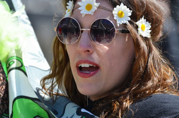 a woman with a head band made of rope and fake daisies, wears round sunglasses, mouth with red lipstick, partially open and talking,