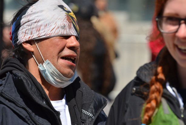 Indigenous man with cap on his head and medical mask under his chin, makes a face, a red head woman in on the right, partially cropped out and out of focus, she is laughing
