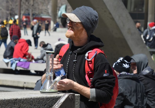 a young man with black hoodie, stands outside at Nathan Phillips square with a bong in his hand, smiling, 420 day event