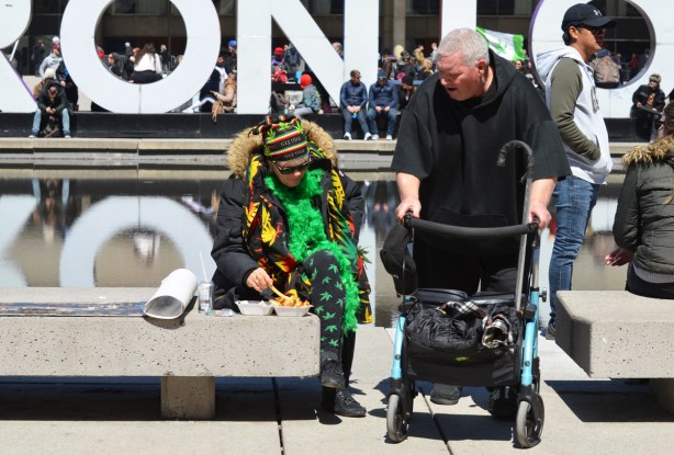 a woman in green and clothes with marijuana leaves on them, sits on a bench at Nathan Phillips, a man in a walker is beside her and talking to her.
