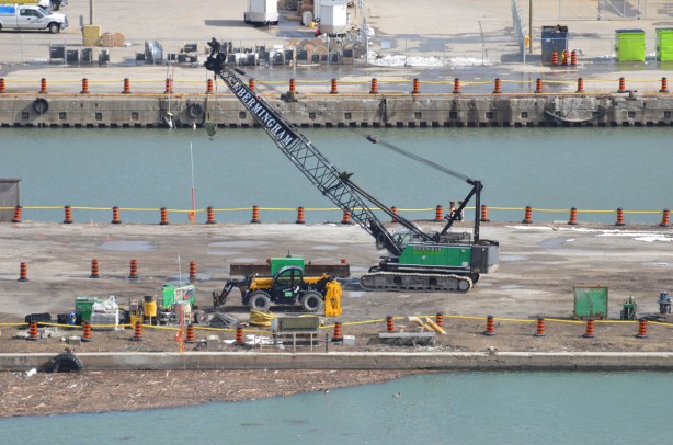 a mobile crane on tractor wheels, green body, is helping dig a hole in the ground on a flat piece of land that has water on two sides. lots of orange and black construction cones around the piece of land