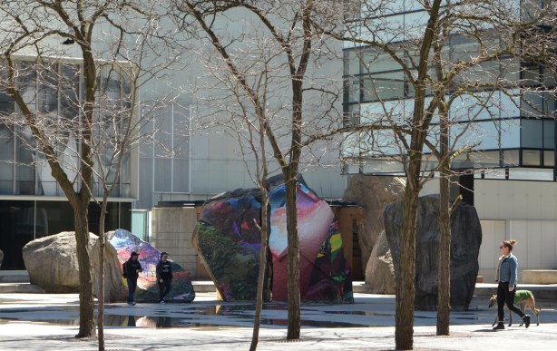 Devonian Square in Toronto, large open area with wading pool (empty at the moment) and large boulders, small trees growing around the edge of the pool, two people walking through the pool area, a woman walking her dog on the sidewalk beside, rocks covered with artwork