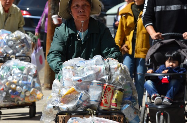 An older Chinese woman is pushing a cart with a large clear plastic bag full of empty aluminium cans, a Chinese man with a similar cart is behind her, other people out walking on the sidewalk