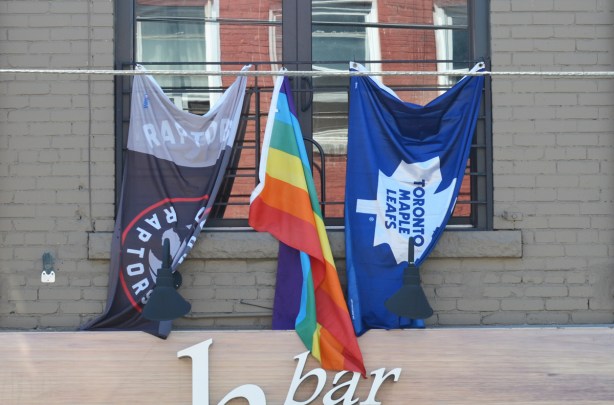 three flags hanging outside a window, a Raptos flag (Toronto basketball) and a Maple Leafs flag (Toronto hockey) as well as a rainbow pride flag