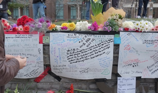 white bristol board taped to a stone wall, condolences and other heartfelt messages written on them, flowers laid across the top of the wall