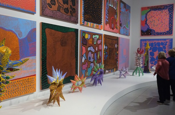 a wall of bright lively paintings by Yayoi Kusama on the wall of the Art Gallery of Ontario. Some women are standing nearby, looking at the paintings. 