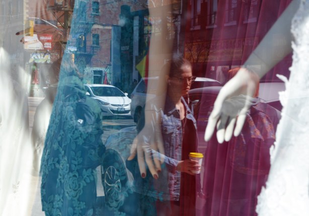 a window with mannequins in long dresses, teal and magenta fabric, reflections of woman passing by with a coffee cup in her hand