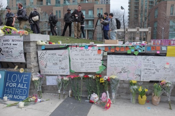 white bristol board taped to a stone wall, condolences and other heartfelt messages written on them, flowers laid across the top of the memorial wall 