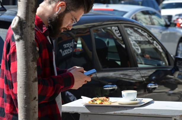 a man outside, standing on sidewalk, with a plate of food and a cup of coffee,, with his phone in his hands 