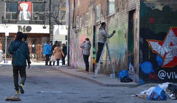 street artist skam is painting text graffiti (his name, tag) in greens and blues on a wall in Graffiti Alley