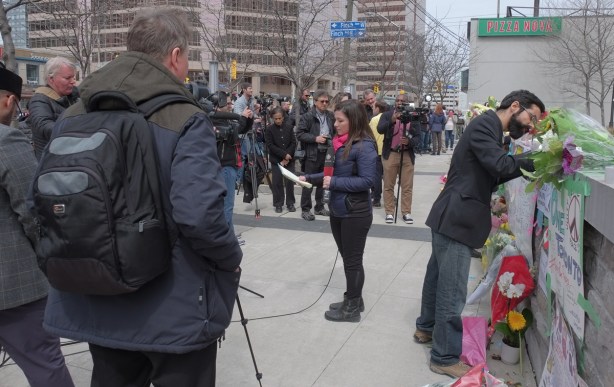 white bristol board taped to a stone wall, concolences and other heartfelt messages written on them, flowers laid across the top of the wall , people, reporters, and photographers standing in front, a man is writing a message 