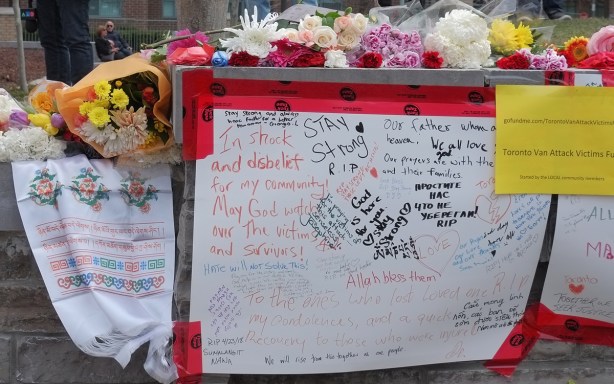 white bristol board taped to a stone wall, condolences and other heartfelt messages written on them, flowers laid across the top of the wall 