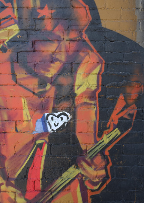 part of mural of a guitar playing in shades of orange and red. A white piece of paper has been stuck on his chest, with a black smiley heart on it. 