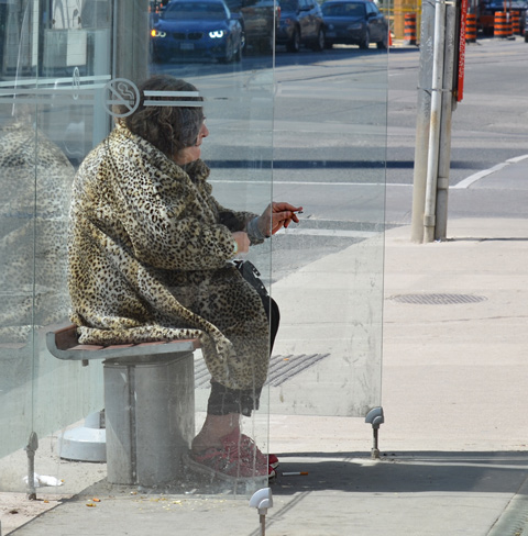 an older woman in pink running shoes and leopard print fuzzy jacket is sitting in a bus shelter, smoking a cigarette, 