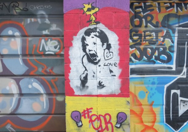 street art on garages, a small yellow Woodstock cartoon character with black sunglasses, on top of a black drawing on white of a girl screaming, a little smiley heart and the word love is beside her ear. 
