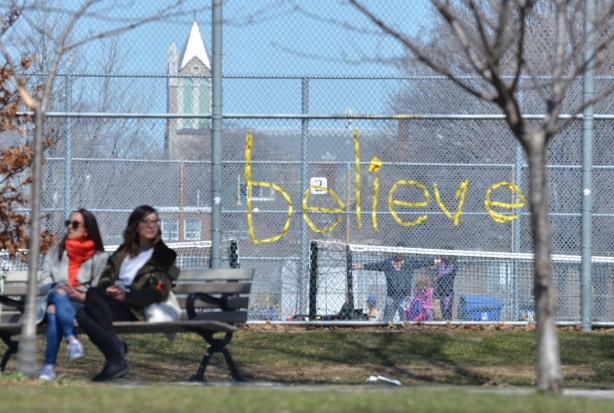the word believe is written in yellow ribbon that has been woven into a chainlink fence around a tennis court, two women on a bench to the side of it, park, 