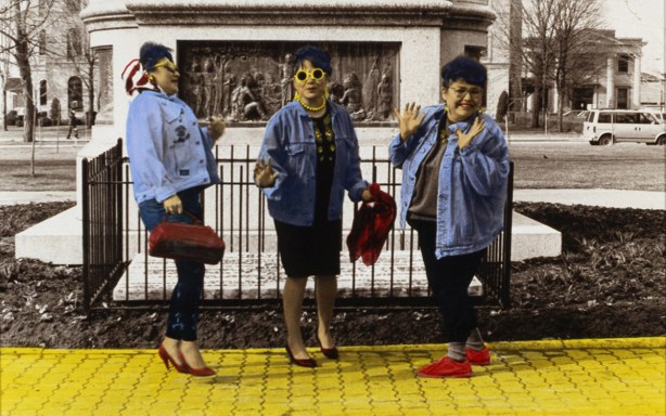 a colourized black and white photo of three women hamming it up for the camera. All wearing red shoes and walking on a yellow sidewalk, beside a metal fence. by Shelley Niro 
