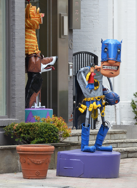 a metal junk sculpture by Patrick Amiot, outside, by the front door of an art gallery, batman, with a very small robin in one of his hands. 