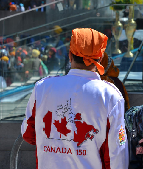 the back of a jacket being worn by a young man, white jacket with red map of canada and the words Canada 150, wearing an orange head scarf tied at the back. 