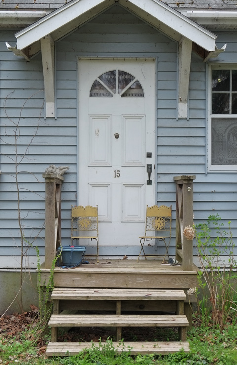 grey wood siding on house with white door and small porch. Two yellow and metal chairs on the porch 