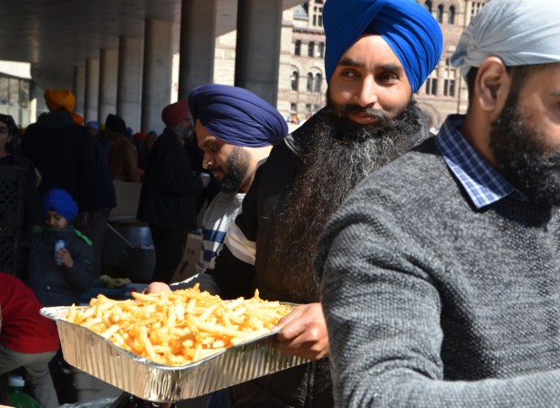 a man in a blue turban and with a long black beard carries a tray of french fries to serve to people 