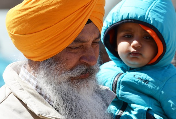 an older sikh man with a long grey beard and an orange turban holds a younger boy in a blue jacket and hood 