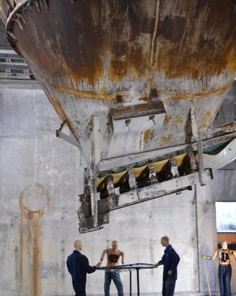 mannequin automatons as part of an art installation in the old Unilever soap factory, concrete floor and walls - three of them stand around a net on a circular frame, a large industrial funnel above them. 