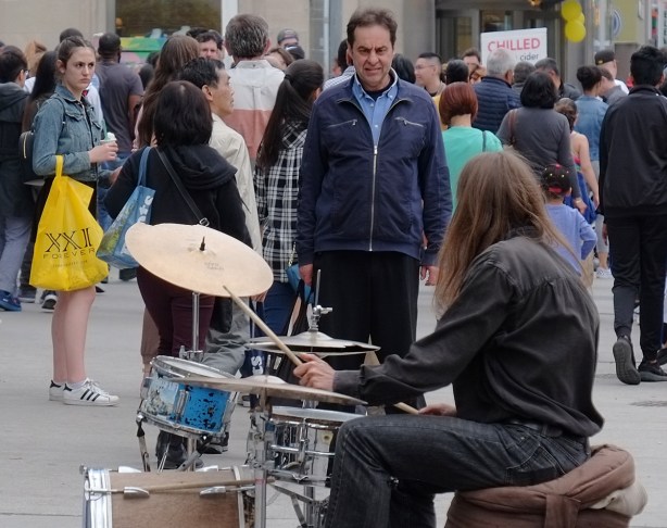a man stands and watches another man playing the drums outside on Yonge street, some other people standing around too 