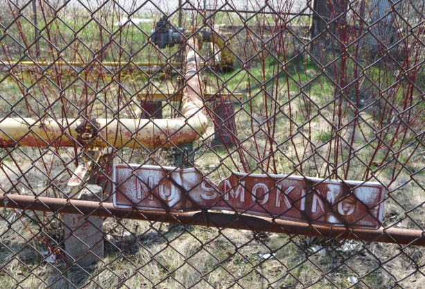 chainlink fence in front of area with yellow pipes, overgrown with weeds and shrubs, sign that says no smoking, white on red, chipped in one corner 