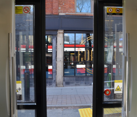 looking out the open doors of a TTC streetcar, as they start to close, see reflection of the streetcar in the window of the store beside the streetcar